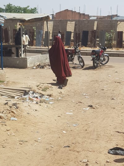 A medium shot depicts a street scene in Dabawa, Nigeria. A man in a long, red robe is walking. Two motorcycles are parked on the side of the road, adjacent to buildings in various states of repair. The ground is sandy and strewn with litter.