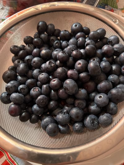 The image shows a close-up, overhead view of a metal colander filled with blueberries. The blueberries are dark blue, plump, and appear to be fresh. They are packed tightly together, filling the colander. The colander has a fine mesh and a silver rim. The background is blurred, but suggests a kitchen or dining setting. There is no indication of the location in the city of Constanța, Romania, or any people, actions, or interactions.