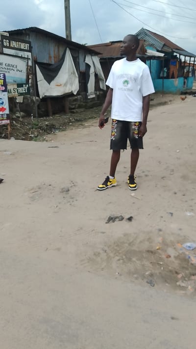 A young man stands on a dirt road in Agbarho, Nigeria. He is wearing a white t-shirt with the text "FORGIVENES" and an emblem that looks like a laurel wreath above a circle. He is also wearing black shorts with colorful embroidered designs and yellow and black sneakers. The man is holding a smartphone in his right hand. In the background, there are several corrugated metal shacks, one of which has a sign that reads "GG APARTMENT". The ground is covered in dirt and some scattered debris, including plastic bags and what appear to be pieces of colored paper. The sky is overcast.