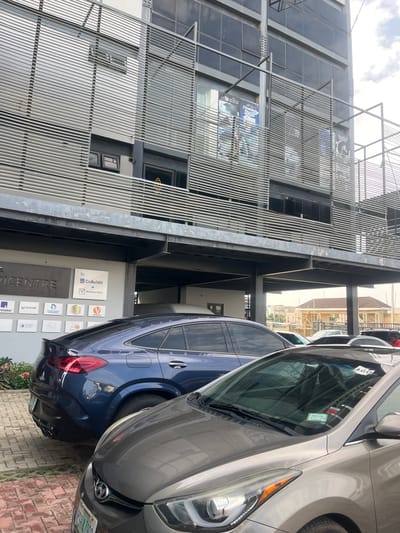 The image shows a street-level view of a modern, multi-story building with a metallic facade and horizontal louvers. Several cars are parked in front of the building. In the foreground, a grey Hyundai Elantra is parked, and to its left, a dark blue Mercedes-Benz GLE Coupe is visible. The building appears to be a commercial or office space, as indicated by various logos and signage on a wall near the entrance, including "EPICENTRE," "CoBuildit + Ventures Park," and other company logos. One of the signs mentions "ABUJA," suggesting the location is in Abuja, Nigeria. In the background, other buildings and a construction site are visible, indicating an urban environment. The sky is cloudy. The image is taken from a slightly elevated perspective, looking towards the building.