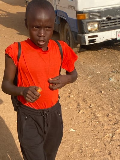 An image taken in Udu, Nigeria, depicts a young male individual outdoors, facing the camera directly with a serious expression and mouth slightly open. He possesses dark skin and short, dark hair, and wears an orange short-sleeved t-shirt, black sweatpants with a drawstring, and a black backpack. In his left hand, he holds a yellow and orange object, possibly a candy or small toy, which features a cartoon-like face. His right hand is positioned near his chest, with fingers slightly curled. In the background, partially obscured by the individual, is a white and blue truck, identified by "DYNA 100" lettering, visible on its side. The truck's front grille, a single headlight, and part of its front bumper with a partially visible license plate are discernible. The ground is unpaved, consisting of light brown dirt or dried earth with scattered small rocks and debris. The scene is illuminated by bright daylight, casting clear shadows to the subject's left.