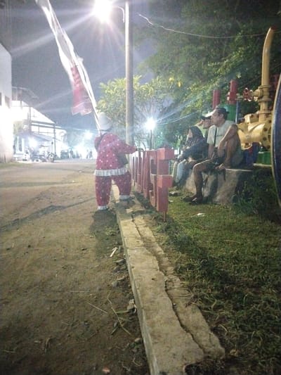 The image depicts an outdoor night scene in what appears to be a public area or park in Sragen, Indonesia.
In the foreground, a person in a whimsical, red and white polka-dotted costume with ruffled white trim and a light-colored hat stands with their back to the viewer. They are holding a long pole from which a large white and red flag, possibly the Indonesian flag, extends upwards towards a bright street light. The costumed figure is positioned on a dirt path next to a concrete curb, appearing to be stepping onto or observing something behind the curb.
To the right, several individuals are seated on a low concrete structure. One man in a light-colored t-shirt and shorts sits casually, while another person wearing a dark head covering is also visible. Behind them, a large, yellow, industrial-themed sculpture with prominent red valves adds to the decorative elements of the area. Large, red, block letters forming a sign are partially visible in front of these seated people, clearly showing "SRA" and hinting at "SRAGEN" or a related word.
The scene is illuminated by multiple artificial lights, including a bright street lamp that casts a strong flare. The background features more buildings with lights, some parked motorcycles, and various trees and foliage. The ground consists of a dirt path on the left and foreground, bordered by a concrete curb, leading to a grassy area on the right. The overall atmosphere suggests a relaxed public gathering or an event at night.