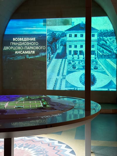 An interior shot captures a museum exhibit focused on the "Construction of a Grand Palace-Park Ensemble." The exhibit features a large, illuminated screen displaying historical images and text in Russian. The text reads "ВОЗВЕДЕНИЕ ГРАНДИОЗНОГО ДВОРЦОВО-ПАРКОВОГО АНСАМБЛЯ," translating to "Construction of a Grand Palace-Park Ensemble." The screen shows aerial views of a palace and formal gardens, as well as a detailed drawing of a Baroque-style building and its surrounding landscape.
In the foreground, a circular, glass-topped display table showcases a detailed architectural model of a palace complex with manicured gardens and waterways. The table's surface reflects the projected images from the screen. Along the edge of the table, a chronological inscription is visible, partially obscured, with the visible dates "1742-1800s." The floor beneath the table features a geometric mosaic pattern. The overall lighting is dim, with the illuminated screen and model being the primary light sources, creating a dramatic and informative presentation. The scene appears to be set within an arched architectural space.
The exhibit is located in the museum of the Arkhangelskoye estate, situated in the dacha settlement of "Arkhangelskoye," Moscow Oblast, Russia.