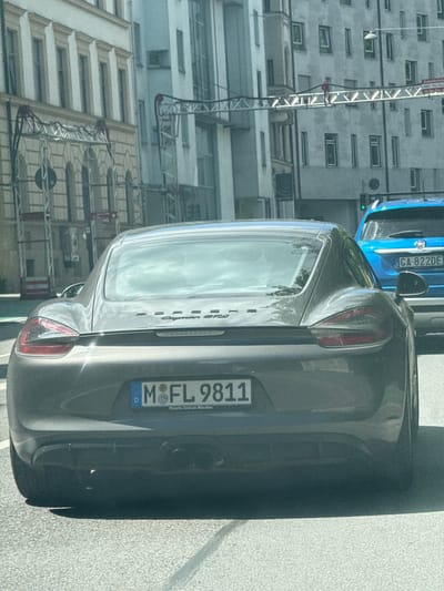 In Munich, Germany, a gray Porsche Cayman S is viewed from the rear, with its license plate reading "M FL 9811." The car is in motion on a street with other vehicles, including a blue Fiat SUV behind it, whose license plate reads "GA 822DE." The background features buildings with architectural details and overhead structures.