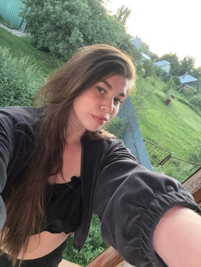A woman with long brown hair is taking a selfie outdoors. She is wearing a black top and a black zippered jacket. The background is a grassy hillside with several small houses and trees. The sky is bright, suggesting it is daytime. The location is a rural area, likely in or near Мещёры, Russia, given the presence of traditional wooden houses and a natural landscape.