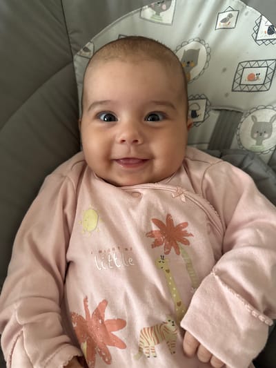 A close-up, high-angle medium shot features a smiling baby girl with dark eyes and dark hair, wearing a pink long-sleeved onesie adorned with cartoon animals and the text "I MIGHT BE little". She is lying on a grey baby bouncer with a patterned fabric backrest featuring animal illustrations. The lighting is natural and bright, casting soft shadows. The baby is looking directly at the camera with a slight smile. The scene depicts a domestic indoor environment, possibly a living room. There is no indication of the location being in Oldham, United Kingdom.