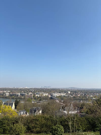 The image presents a panoramic elevated view overlooking the city of Le Pecq, France, under a clear, bright blue sky. The foreground features dense green foliage, including trees with bare branches and some vibrant green bushes, several displaying bright yellow blossoms.

The midground is dominated by an urban landscape with a winding river, identifiable as the Seine, flowing horizontally from left to right. Along the riverbanks, a mix of residential buildings is visible, including multi-story structures with light-colored facades and traditional houses with gray slate roofs. A prominent bridge with several arches spans the river, carrying vehicular traffic.

In the background, a sprawling cityscape extends into the distance, characterized by a mix of residential and commercial buildings interspersed with significant green spaces. A distinct cluster of high-rise skyscrapers, indicative of the La Défense business district, is clearly visible on the horizon towards the right-center. Further to the left on the horizon, another, less defined, urban cluster suggests the broader Parisian metropolitan area. No individuals, specific actions, or interactions are discernible. The scene is illuminated by bright daylight, indicating clear weather.