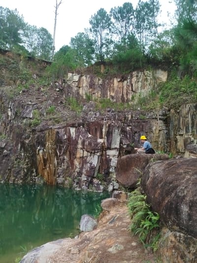 A person wearing a yellow hard hat and a blue shirt sits on a large rock by the edge of a quarry with green water. The quarry walls are steep and made of layered rock formations, with some vegetation growing on them. Pine trees are visible on the ridge above the quarry. The sky is overcast.