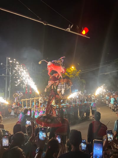 This image captures a nighttime street scene during a ceremonial procession in Munggu, Indonesia. The central focus is a large, multi-tiered effigy, likely an Ogoh-Ogoh, being carried by approximately twenty men. The effigy's uppermost section depicts a figure, possibly a deity, dressed in red and purple attire, riding a light pink bull-like creature. Below this, two additional figures are integrated into the effigy structure; one figure is notably wearing a black and white checkered sarong.
Bright sparks from multiple pyrotechnic devices, resembling large sparklers, erupt horizontally from both sides of the effigy's base, creating plumes of smoke that partially obscure the background. The effigy is supported by a robust bamboo and wooden framework, which is shouldered by the uniformed carriers. These individuals wear matching blue-green t-shirts with patterned sleeves and traditional sarongs.
A dense crowd lines the street on both sides, observing the procession. In the foreground, numerous spectators are visible from behind, many holding smartphones vertically to record the event. Some foreground individuals wear dark shirts, one with visible text "KUMANDA." An individual near the lower center holds a traditional red and white Balinese ceremonial umbrella. Overhead, a traffic light displays a solid red signal, and utility wires crisscross the dark sky. Streetlights illuminate the scene, casting light on palm trees and distant buildings.