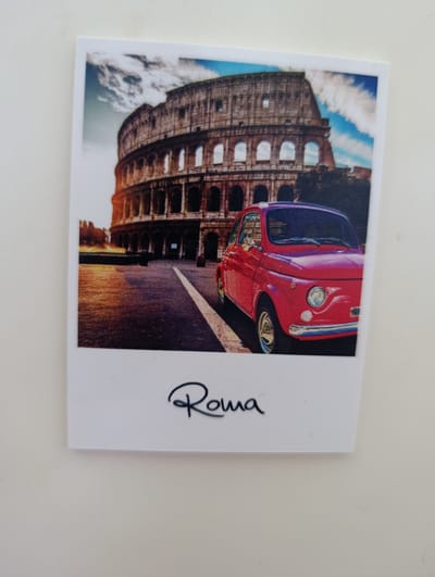 The image is a photograph of a postcard featuring the Colosseum in Rome, Italy, with a red Fiat 500 car parked in front of it. The Colosseum is depicted in the background, and the sky is visible with clouds. The red car is in the foreground, angled towards the viewer. Below the image, the word "Roma" is written in a cursive font. There is no indication that this image is related to the city of Razgrad, Bulgaria.