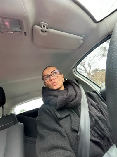 A person is depicted seated in the front passenger seat of a vehicle in Saint-Cyprien, France. The individual, appearing to be female, has light-toned skin, dark hair pulled back, and wears rectangular tortoiseshell-patterned glasses and a gold-colored hoop earring on the visible right ear. Her lips are pursed in a pout, and her gaze is directed upwards and to the left. She is dressed in a dark, textured coat and a voluminous, dark, chunky knit scarf wrapped around her neck. A light-colored seatbelt diagonally crosses her chest. The car's interior features a light gray headliner with an overhead light and sun visor. Through the window on the right, raindrops are visible on the glass, and bare tree branches are discernible in the blurred background under an overcast sky, indicating rainy or recently wet conditions outdoors. The top of another car seat is visible on the far left.