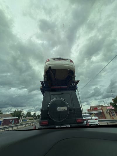 The image, taken from inside a car, shows a black Mercedes-Benz G-Class towing a white Volkswagen Golf in Troitskoye, Russia. The Volkswagen is elevated and attached to a specialized tow bar or lift on the back of the Mercedes. The background reveals a cloudy sky. There is also a building with a shop that says "ХозМаркет". The photograph captures a street scene in the specified location, emphasizing the unusual method of vehicle transport.