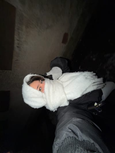 A person stands in a dimly lit outdoor space, likely a yard or alley. They wear a thick, dark jacket and a large, fluffy white scarf wrapped around their head and neck, partially obscuring their face, though one eye is visible. Gloved hands extend, the left gesturing downward. The background shows a dark and grainy wall, possibly concrete, with some uneven textures and variations in light. A hint of a small brick is visible. The overall image is tilted, adding to the disorienting and slightly eerie feel.