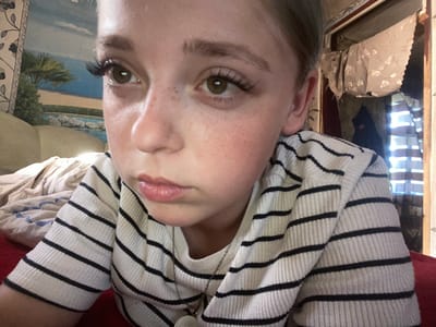 This is a close-up, overhead, upside-down portrait of a young person. The subject is wearing a cream-colored sweater with horizontal black stripes and a delicate silver chain necklace. Their face is turned towards the camera, with freckles scattered across their nose and cheeks. They have large, expressive brown eyes, one of which has dramatic false eyelashes. The person's lips are slightly parted, appearing to be glossy. The background is blurred and indistinct, but hints of a patterned fabric and what appears to be a tapestry or framed picture with a beach scene are visible. The lighting suggests an indoor setting, possibly during the day due to the bright light coming from a window on the left side of the frame. The orientation of the photo implies it was taken by the subject themselves, looking down at their phone or camera. The overall impression is one of youthful self-expression and a casual moment captured.