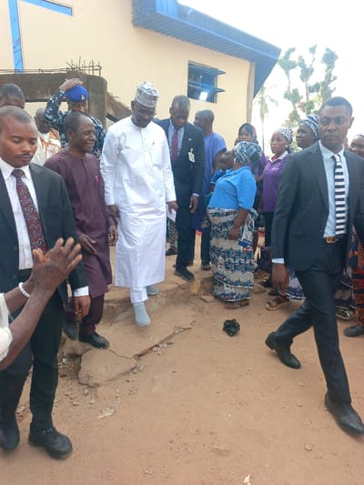 The scene depicts an outdoor gathering in Ube, Nigeria, with approximately fifteen to twenty individuals present on a reddish-brown dirt path in front of a light-colored building.
In the central mid-ground, a prominent male figure wearing a white traditional agbada-style garment and a white cap is stepping down from a slight elevated concrete section onto the dirt path. He wears light grey socks and glasses, looking downwards. To his left, a man in a maroon traditional top and dark pants smiles, looking towards the right. To the right of the central figure, a man in a dark suit with a red tie and a visible badge is also descending, looking down. Behind these individuals, a police officer in a dark blue uniform and cap is partially visible, adjusting his cap.
On the far right in the foreground, a man in a dark suit with a black and white striped tie walks towards the right, looking straight ahead. His belt features a gold-colored buckle. In the mid-ground right, a woman in a light blue top and a patterned wrapper skirt stands on the dirt path, looking downwards. A child is partially visible on her back, obscured by her clothing. Several other women in patterned attire and head coverings are visible behind her.
On the far left in the foreground, a man in a black suit with a patterned tie is partially visible, facing slightly right. Adjacent to him, an older pair of hands adorned with a white beaded bracelet is clapping.
The ground consists of an uneven, unpaved reddish-brown dirt surface with visible cracks and small pieces of debris. A dark, irregularly shaped object rests on the path in the mid-ground. The background features a light beige building with at least two windows with blue frames on an upper level, topped by a blue corrugated metal roof overhang. Exposed concrete and rebar structures are visible near the building on the left side. The scene is illuminated by natural daylight.