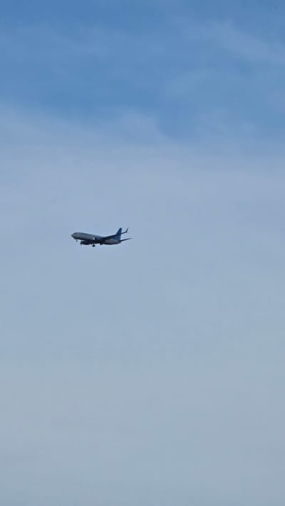 A wide shot captures a blue-grey commercial airplane in mid-flight against a pale blue sky with wispy clouds. The aircraft, appearing to be descending, is positioned slightly left of center in the frame. Its landing gear is extended and its propellers are visible. The airplane is the sole subject in the vast expanse of the sky. The image does not provide any information indicating its location in Bulgakovo, Russia.