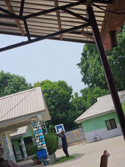 The image depicts an outdoor scene in Unguwar Rimi, Nigeria, possibly a schoolyard. The angle is from a low vantage point, looking up between structures.
On the left, a section of a corrugated metal roof supported by a dark brown metal framework is visible. To the right, there's a two-story building painted in shades of green and grey, with a white air conditioning unit mounted on the exterior wall. Adjacent to the building is a basketball court with a man in dark clothing standing beneath the basketball hoop, seemingly engaged in an activity. A blue water dispenser sits near a short wall decorated with blue and white geometric patterns. Lush green trees fill the space between the buildings, partially obscuring the sky.