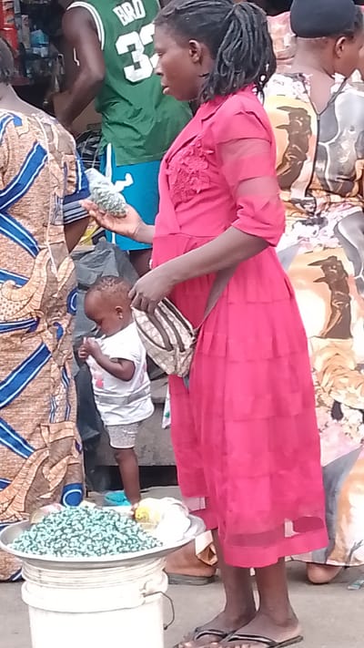 In Agbarho, Nigeria, a woman in a bright pink dress stands to the right, holding a small bag of light green pebbles. In front of her, a young child in a white t-shirt and grey shorts looks down, holding something small in their hands. To the left, another person wears a patterned dress with blue and tan accents. Behind them, a person in a green basketball jersey with the number "32" is visible. In the foreground, a metal tray filled with a large quantity of the light green pebbles sits atop a white bucket. The scene appears to be an outdoor market.