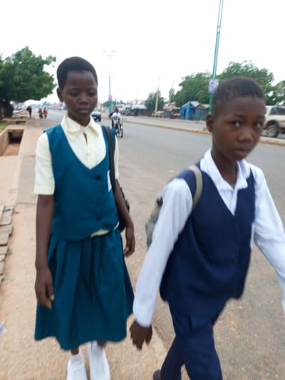In Garko, Nigeria, two children, a girl and a boy, walk along a dirt sidewalk adjacent to a paved road. The girl, in the foreground and to the left, wears a light yellow collared shirt and a dark teal sleeveless vest with two buttons. She also has a matching pleated skirt and white sneakers. A dark bag is slung over her left shoulder. Her gaze is directed forward. The boy, to the right and slightly behind, wears a white long-sleeved shirt and a dark blue sleeveless vest, paired with dark blue pants and dark shoes. A light-colored backpack is visible on his back. He is also looking forward.

The road is a two-lane asphalt surface. Traffic on the road includes a motorcycle with a rider in the background, and a light-colored SUV further down the road. The environment includes sparse trees and buildings in the distance. The sky is overcast.
