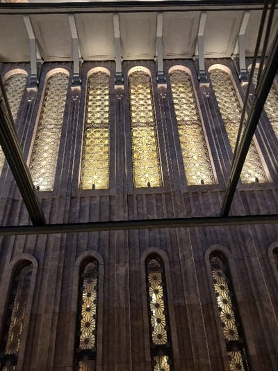 The image shows a low-angle view of a building's facade with a series of tall, narrow windows arranged vertically. The windows are framed by dark stonework, with an intricate geometric pattern etched into the glass. The building is constructed of light gray stone and is located in Istanbul, Türkiye, at the coordinates (41.037, 28.984). It is likely to be a historic or religious building. The light from inside the building illuminates the windows, creating a warm glow. There are no people visible in the image.