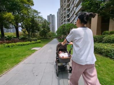 A woman is pushing a baby in a stroller on a paved path in Shunde District, China. The path is bordered by green grass and trees. High-rise buildings are visible in the background. The woman is wearing a white t-shirt and pink pants. The baby is lying in the stroller.