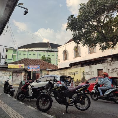 The image depicts an urban street scene in Sukabumi, Indonesia, with GOR Sukabumi (Gelanggang Olahraga Sukabumi) visible in the background. The main GOR building is large, green, multi-storied, and features a prominent dark domed roof. To the right of the GOR, a white building with several arched, light-colored windows is visible, partially obscured by a large, leafy tree.
In the mid-ground, a row of smaller, single-story buildings with tiled roofs and corrugated metal shutters line the street. One shop has a "SWADAYA" sign, and another displays a "PANGKAS RAMBUT" (haircut shop) sign. A low concrete wall topped with barbed wire runs in front of the white building and extends along the street.
The foreground features a paved sidewalk and street. Four motorcycles are parked on the sidewalk/shoulder: a black scooter on the far left, a red scooter, a prominent black and yellow touring motorcycle on a kickstand, and another dark scooter. A white multi-purpose vehicle (MPV) is parked behind these motorcycles. On the street, a person wearing a light blue jacket and an orange helmet is riding a red and white scooter, moving from right to left, passing a parked red vehicle. The sky is partly cloudy, and the scene is illuminated by natural daylight, casting some shadows. Overhead utility lines and a street lamp are visible in the upper left.
