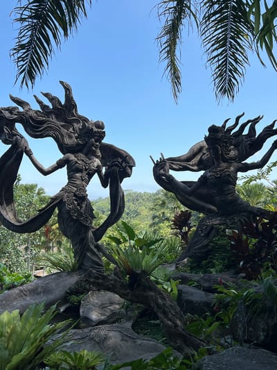 The image shows two Balinese statues amidst lush greenery and a clear blue sky. The statues are made of dark stone or metal. The lower statue is a female figure with long flowing hair, adorned with intricate jewelry and flowing garments. The upper statue is a male figure with a fiery headdress and expressive gestures. They appear to be reaching for each other. Palm fronds frame the left side of the image. The background reveals a landscape of rolling hills covered in dense vegetation. The location is Kedewatan, Indonesia.