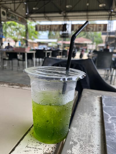 The image captures a green beverage in a clear plastic cup with a black straw, placed on a weathered, gray surface. Condensation droplets cling to the cup's exterior. The background reveals an outdoor setting, possibly a cafe or open-air establishment in Seuneubok Teungoh, Indonesia, as indicated by the urban environment with metal structures, buildings, and blurred figures and vehicles. The image is slightly rotated.