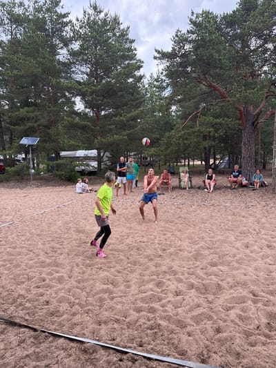 In Zelenaya Roshcha, Russia, a sandy beach volleyball court is set up amidst a pine forest. A group of people are playing volleyball, with one person in the foreground in a neon green shirt and black leggings, and another in blue shorts hitting the ball. Spectators are seated on chairs and the sand in the background, observing the game. Beyond the court, trees and campers are visible, suggesting a recreational area within the forest. The sky is overcast.