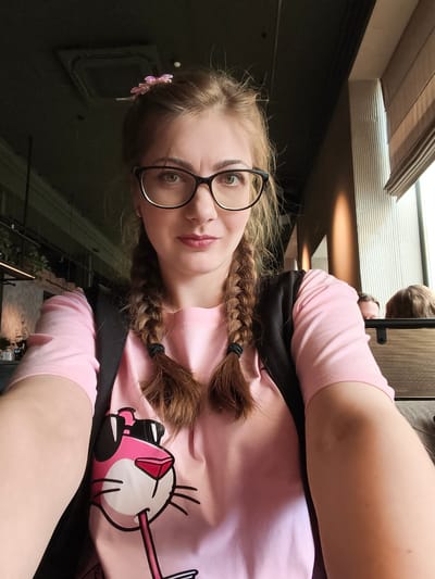 A young woman with blonde hair in two braids and wearing glasses and a pink t-shirt with a Pink Panther graphic is taking a selfie in what appears to be a cafe or restaurant in Voronezh, Russia. The background shows interior elements such as a ceiling, lighting fixtures, and a window with blinds. Two other individuals are partially visible in the background. The lighting suggests it is daytime.