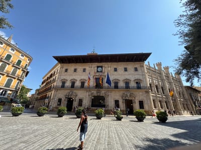 The image captures a wide-angle view of the Plaça de Cort in Palma de Mallorca, Spain, under a clear blue sky. The foreground features a cobblestone plaza with several large, dark planters containing lush green bushes. A woman with long dark hair, wearing a black tank top and light blue shorts, is walking away from the camera towards the center of the plaza.
Dominating the background is the Palau de l'Ajuntament de Palma (Palma City Hall), a grand sandstone building with a central clock and adorned with flags. To the left, a bright yellow building with green shutters and balconies partially frames the scene. The architecture is characteristic of the historic city center, with intricate details on the buildings and a sense of old-world charm. Several other people are visible in the background, walking on the plaza and in front of the buildings, suggesting a busy public space. Trees with green foliage are present on the left and right edges of the frame, adding a touch of nature to the urban setting. Sunlight casts sharp shadows across the plaza, indicating a bright, sunny day.