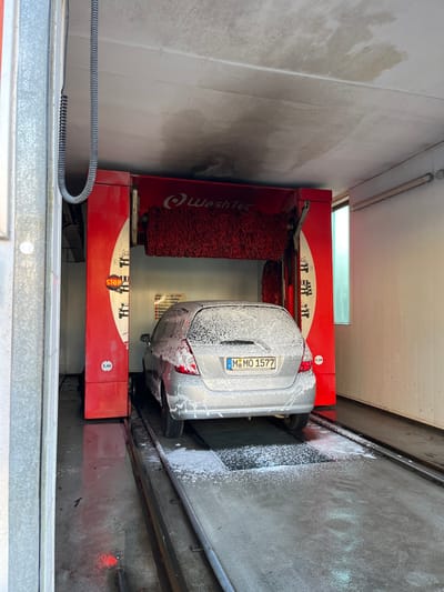 A silver hatchback vehicle, possibly a Honda Fit/Jazz, is situated within an active, automated car wash bay in Meisenheim, Germany. The car is coated in a thick layer of white soap foam, indicating it is undergoing a wash cycle. Its German license plate displays "M MO 1577". The vehicle is positioned on metal guide rails embedded in a wet, dark concrete floor, with a gridded drainage area directly beneath its rear tires.

The car wash mechanism, branded "WashTec," is prominently red with white accents and features large, cylindrical red brushes positioned above and behind the car. A circular red "STOP" sign is visible on the left pillar of the machinery, alongside a white label indicating "2,40" (likely a height limit in meters). Additional German text is present on a partially obscured sign on the right pillar of the machine. The interior environment consists of white walls, with a tall, narrow window on the right allowing ambient light into the bay. The concrete ceiling above is stained with dark, irregular patches, suggesting water exposure or discoloration over time. A grey, coiled cable hangs vertically from the ceiling on the left side of the frame. No individuals are present in the scene.