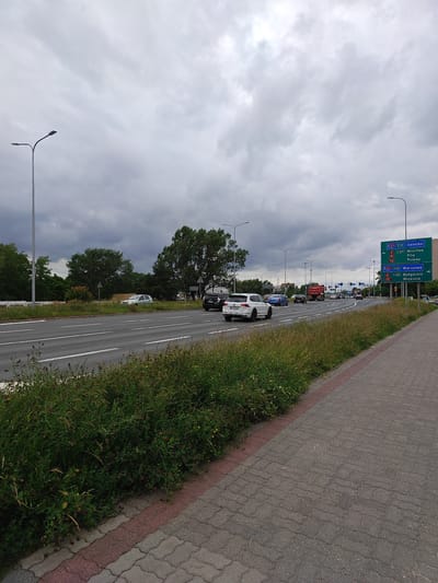 This is a wide shot of a road in Poznań, Poland, under a cloudy sky. A multi-lane road runs diagonally across the frame from left to right, with cars and trucks driving on it. The road is bordered by a grassy median on the left and a paved sidewalk on the right. A street light stands on the median to the left of the frame. On the right side of the road, a green directional sign points towards multiple cities, including Świecko, Wrocław, Piła, and Warsaw. The sign also indicates the A2 and E30 highways. The overall scene depicts a typical urban roadside environment.