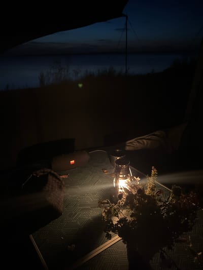 An outdoor camping scene at dusk or night near Am Salzhaff, Germany. The foreground is dominated by a table covered with a patterned cloth, illuminated by a vintage oil lantern. A bouquet of dried flowers is placed next to the lantern. To the left, a dark fabric object and a portable speaker with an illuminated orange light are visible. In the mid-ground, faint outlines of tents or shelters are present. The background shows a dark body of water under a twilight sky, with silhouetted vegetation along the shoreline.