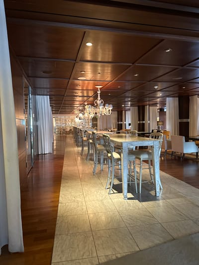 The image depicts a long, narrow dining area with marble-topped tables and white metal chairs. Chandeliers hang along the length of the room. A dark wood paneled wall with embedded lighting is on one side, while a stone-tiled floor is on the other. At the end of the tables, white curtains are hung. The space appears to be in a hotel or upscale event venue.