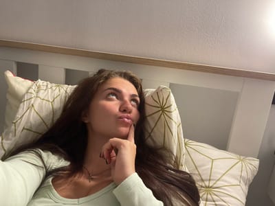The image shows a young woman lying in bed, looking upwards with her finger on her chin in a pensive pose. She has long, dark hair and is wearing a light green ribbed top and a silver cross necklace. The bed has white pillows with a geometric pattern of green lines on a white background. The headboard of the bed is white with a wooden trim. The ceiling is textured and appears to be painted white. The lighting suggests an indoor setting, likely a bedroom, during the evening or night. There is no information in the image to indicate the location in the city of Мосметаллоконструкция Онт, Russia.