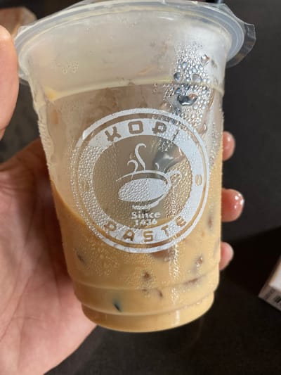 A close-up, horizontal shot shows a person's hand holding a clear plastic cup filled with an iced, light brown beverage, likely a milky coffee. The cup is wet with condensation, indicating a cold drink in a warm or humid environment. Several fingers are visible gripping the cup, with a thumb supporting it from below.
The cup features a white, circular logo prominently displayed on its side. The logo reads "KOPI" on the left and "PASTE" on the right, encircling a stylized image of a coffee cup with steam rising from it. Below the coffee cup icon, smaller text reads "since 1986". Ice cubes are visible within the beverage, which has a smooth, creamy appearance. The background is dark and out of focus, providing no specific details about the immediate surroundings beyond suggesting an indoor setting. The image captures the act of holding a refreshing drink, possibly consumed in Tanjongkapai, Indonesia.