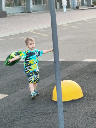The image shows a young boy standing on an asphalt surface, likely a playground or park, given the presence of a yellow dome-shaped object and a pole. The boy is wearing a brightly colored t-shirt and shorts with a racing-themed pattern. He is holding a green, somewhat deflated balloon with a string in his left hand and the string of another balloon in his right hand, which extends towards the pole. His expression is somewhat serious or thoughtful. The setting appears to be outdoors, with a building visible in the background. The lighting suggests it might be late afternoon or early evening, with the sun casting shadows. The ground is a textured asphalt. Text on the boy's clothing includes "Super," "Sporting," and what appears to be the number "10," along with images of cars and trucks. The overall scene conveys a moment of childhood play.