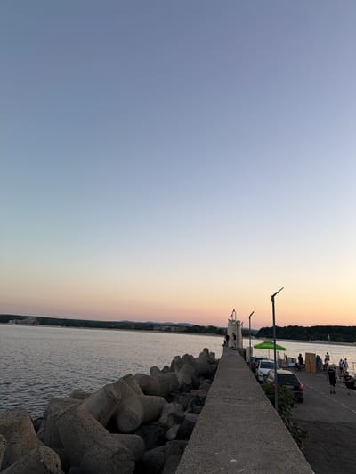 This media file captures a seaside scene at dusk in Primorsko, Bulgaria. The image is oriented vertically, with the sky occupying the majority of the frame. The sky displays a gradient of soft colors, transitioning from a pale blue at the top to warm hues of peach and yellow near the horizon, indicative of sunset.
On the right side of the image, a concrete structure, possibly a breakwater or pier, runs along the edge of the water. Large, concrete tetrapod-shaped blocks are piled on the edge of this structure, designed to break waves. Beyond the concrete structure, a body of water, likely the sea, is visible with gentle ripples.
Further in the background, a shoreline is lined with trees and some buildings. A paved area runs parallel to the concrete structure, where several people are seen. Some individuals are standing or walking, while others are seated. There are also cars parked on this paved area, including a black Audi with a license plate that appears to read "PA 3344 KA". A green umbrella is also visible. A lighthouse-like structure stands near the edge of the paved area.
The overall atmosphere suggests a relaxed evening scene. The soft lighting and the presence of people enjoying the waterfront indicate a peaceful time of day. The weather appears calm.