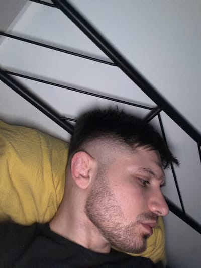 A man is lying on his side on a bed, with a yellow blanket beneath his head. His head is close to the metal frame of the bed. The background is a plain, light-colored wall. The location is not discernible from the image and therefore cannot be placed within the city of Pentre, United Kingdom.
