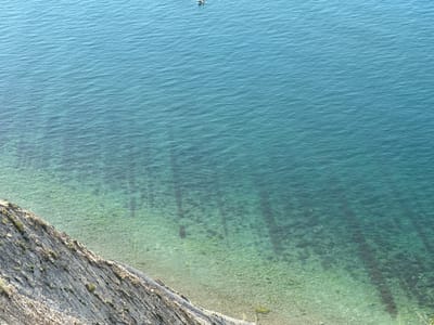 The image shows a view of the sea from a cliff overlooking the coast of Myskhako, Russia. The water is a clear turquoise color, with ripples on the surface. The seabed is visible in the shallower areas, with rocks and seaweed. A small boat with two people is visible in the distance. The cliff face on the left is rocky and sparsely vegetated. The scene depicts a calm day at the seaside.
