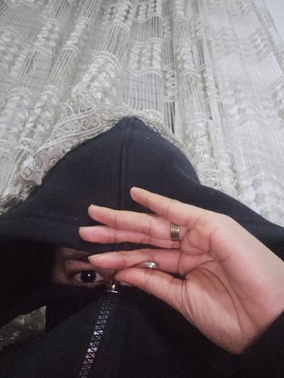 A close-up image reveals a hand with rings on the ring and middle finger. The hand is near a black zipper hoodie. Peeking out from inside the hoodie is a glimpse of dark fur and a single dark eye, suggesting an animal, possibly a dog or cat, is concealed within. A sheer white lace fabric is partially visible behind the hoodie. The black hoodie and lace backdrop create a contrast with the light skin tone of the hand.