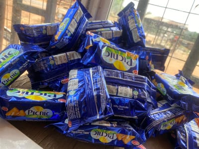 The image displays a substantial, disorganized pile of approximately two dozen "Picnic Milky Shortcake Biscuits" packets. Each packet is enclosed in a glossy blue foil wrapper, prominently featuring the brand name "Picnic" in yellow and green text, accompanied by illustrations of biscuits and a glass of milk. Barcodes and batch numbers, such as "0725", are visible on several packets. One legible label on a wrapper identifies the manufacturer as "MAESTRO FOOD & BEVERAGES LTD. No 1, Boka Lane, Off Lekki-Epe Exp. Way, Ajah, Lagos," confirming a Nigerian origin. The biscuit packets rest upon a light-colored, planar surface, possibly wood or laminate. In the blurred background, an outdoor environment is discernible through a window with a grid-like structure, revealing distant vegetation and undefined structures characteristic of a semi-urban or rural landscape. No individuals are present, and no actions or interactions are depicted. The scene is located in a building within Okura, Nigeria.