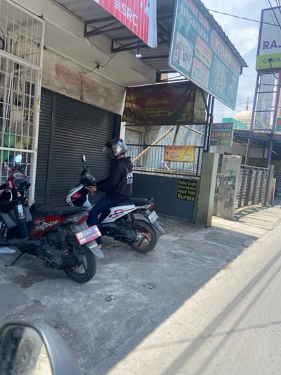 The scene takes place in front of a commercial building on a street in the city of Kecamatan Dayeuhkolot, Indonesia, located at latitude: -6.965, longitude: 107.593. A young male is sitting on a white motorcycle, wearing a black hoodie with a red and white logo, blue jeans, and a white helmet. The motorcycle has a red license plate that reads "6327," which is a local Indonesian license plate. The motorcycle is parked on the sidewalk in front of a building with an awning that has a sign written in Indonesian. The building also has several other signs, some of which have prices and information about the store. The signs are all in Indonesian. The street is paved and grey, with white lines. The building has a closed metal gate. The sidewalk is narrow, and there is some vegetation growing between the sidewalk and the street. The sun is shining, and there is a shadow on the street. The sky is clear and blue. The store sign appears to advertise a Singaparna Kurnia restaurant, which likely serves local Indonesian cuisine.