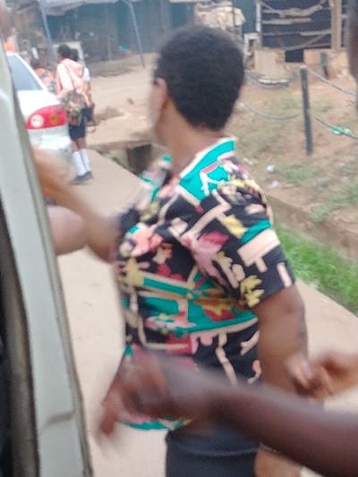 The image portrays an outdoor street scene in Orie Uratta, Nigeria. In the midground, a person with short, dark hair, seen from behind and facing slightly right, wears a short-sleeved shirt patterned with black, teal, pink, yellow, and white geometric and abstract shapes, over dark trousers. Their right arm is partially extended forward. In the lower right foreground, two dark-skinned hands are visible, with one appearing to interact with the main subject's right arm or hand area.
To the left, the side of a white vehicle, possibly a car, is partially visible, displaying a red taillight or reflector. Further in the background, another individual is seen walking away, wearing a light-colored top (possibly peach), dark shorts, and white socks, carrying a patterned backpack. The immediate ground consists of a light-colored, paved surface, likely a sidewalk. To the right of this path, a concrete drainage channel is visible, bordered by green vegetation. The background features blurred structures consistent with an urban or semi-urban environment, including what appears to be corrugated metal and dark poles. The scene is illuminated by ambient daylight.