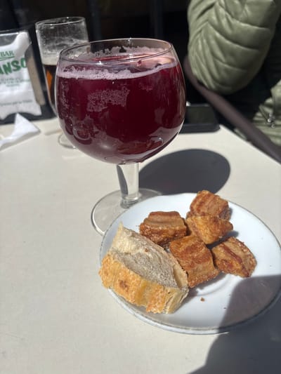 The scene depicts an outdoor table setting at a bar or restaurant in El Espinar, Spain, under direct sunlight. A large, stemmed goblet glass, filled with a dark red, effervescent beverage with surface foam, occupies the central foreground. Adjacent to it on the right, a small white plate holds six pieces of golden-brown, fried pork belly (torreznos) and one slice of rustic bread. Distinct shadows are cast on the light-colored tabletop. In the background, a taller glass with a dark amber liquid is partially visible. To the left, a white paper napkin or menu displays partial green text, including 'BAR' and 'ANSO.' A person wearing an olive-green quilted jacket is seated in the upper right background, partially visible, with a dark rectangular object (possibly a smartphone) on the table nearby.