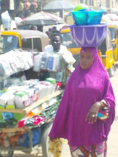 This image captures a bustling outdoor market scene in Fagge D1, Nigeria. In the foreground, a woman dressed in a vibrant purple hijab and matching loose garment, with patterned trousers visible below, walks towards the right. She expertly balances a stack of three plastic bowls on her head: a large purple bowl forms the base, with a blue bowl nested inside it, and a smaller green bowl placed within the blue one. Her right hand, adorned with rings, holds a multi-colored object. To the left of the woman, in the mid-ground, a dark-skinned man wearing a white shirt or cap stands partially obscured by a mobile vendor's cart. He looks towards the viewer. The cart features multiple shelves laden with various packaged goods, including what appear to be phone cases and other small consumer items. In the blurred background, several yellow auto-rickshaws are visible, alongside other market stalls with awnings and numerous indistinct figures, indicating a densely populated commercial area under bright daylight.