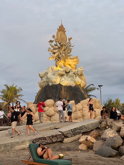 The scene depicts a public gathering area at dusk or early evening in Kerobokan Kelod, Indonesia, under an overcast sky. A large, multi-armed golden statue, possibly a deity, serves as the central focal point, illuminated by spotlights. It stands atop a dark, conical base surrounded by lighter, irregular rock-like formations.
In the foreground, on sandy ground, a person wearing a baseball cap, sunglasses, and a dark top reclines on a green lounge chair, actively looking at a mobile phone. A green coconut drink rests on a small, dark surface next to the chair.
Immediately behind this, a series of broken concrete slabs, marked with graffiti, form part of the terrain leading to the statue. Large, grey and brown rocks are scattered across the sandy ground.
Numerous individuals are present throughout the mid-ground. From left to right:
* A man in a black shirt and khaki shorts walks with a woman in a black dress, both appearing to hold hands.
* Near the base of the statue, several people are standing and conversing. One man in a red shirt and a grey fanny pack is eating a cob of corn.
* A woman in a black swimsuit and white headband stands alone, observing the surroundings.
* Further to the right, multiple individuals are seated on the large rocks, some looking at mobile phones.
In the background, on both the left and right sides, green palm trees are visible. A black street light pole stands on the right. The overall environment suggests a waterfront or beach-adjacent public space with recreational activity.