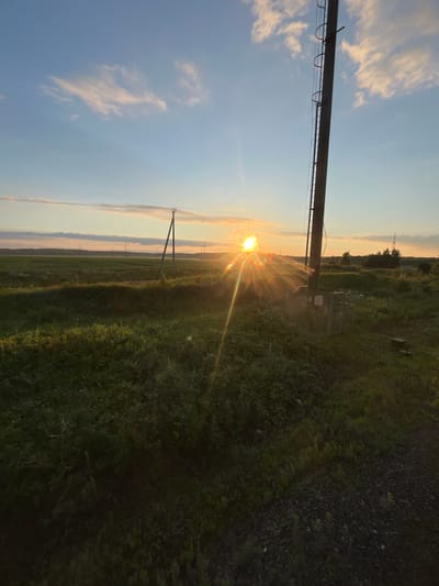 The image depicts a rural landscape at sunset, likely in the vicinity of Kauchuk Stantsiya, Russia. A tall, dark utility pole with a visible ladder dominates the right side of the frame, positioned in a field of green grass and low-lying vegetation. The sun, a bright, radiant orb, is setting near the horizon in the center of the image, casting long rays of light across the sky and landscape. The sky is a gradient of blue and pale orange, with scattered white clouds. In the distance, a line of trees and other utility poles are visible. The ground in the foreground appears to be a mix of gravel and grass. There are no people or animals present in the scene. The overall impression is one of a serene, natural setting bathed in the warm glow of the setting sun.