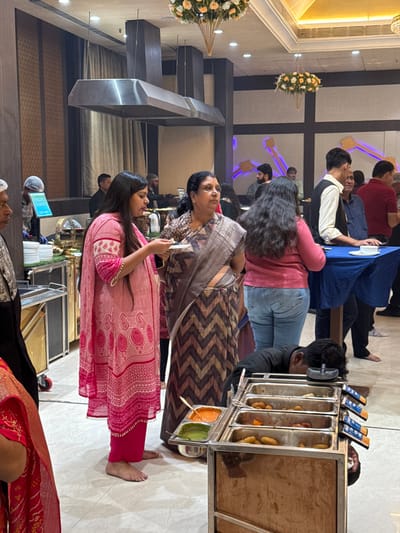 A group of people are inside what looks like a banquet hall. There is a buffet area with several food items. A woman in a pink dress is holding a small white card, while another woman in a patterned sari stands next to her. There is a table with blue tablecloth and seated guests including a person in a red shirt. Decorative floral displays are visible in the background.